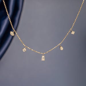 Könnte beinhalten: Eine goldene Halskette mit fünf hängenden Anhängern, die jeweils mit einem klaren Edelstein in unterschiedlicher Form besetzt sind, darunter Marquise-, Rund-, Baguette-, Tropfen- und Quadratschliffe, befestigt an einer feinen, texturierten Goldkette.