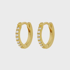 Könnte beinhalten: Ein Paar goldene Creolen mit einer Reihe kleiner, klarer, rund geschliffener Steine, die entlang der vorderen Außenkante jedes Rings eingelassen sind. Die Ohrringe haben eine polierte Metalloberfläche und einen Klappverschluss.