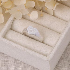 Può includere: Un anello d'argento con una faccia a forma di cuore. L'anello è presentato in un portagioie beige. Il cuore ha una superficie strutturata. Un gioiello semplice ed elegante.