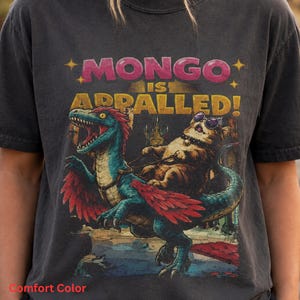 May include: A charcoal gray t-shirt featuring a graphic print of a cat wearing sunglasses riding a blue feathered dinosaur. The text above the illustration reads MONGO IS APPALLED! in pink and yellow block letters. The shirt has a crew neckline.