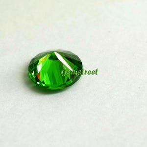 May include: A round, faceted, vibrant green gemstone with a brilliant cut that reflects light. The stone features a polished surface with multiple triangular facets and the word Gemstreet printed in green text across the center of the gemstone.