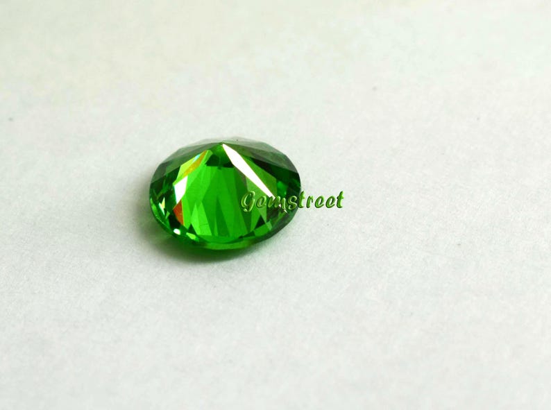 May include: A round, faceted, vibrant green gemstone with a brilliant cut that reflects light. The stone features a polished surface with multiple triangular facets and the word Gemstreet printed in green text across the center of the gemstone.