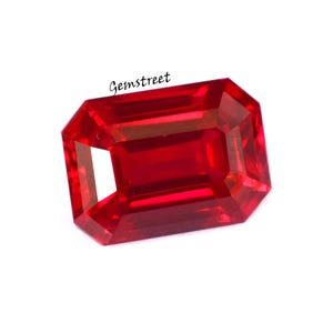 May include: A vibrant, rectangular-cut red gemstone, possibly a ruby, with facets that catch the light. The gemstone is set against a white background. The text "Gemstreet" is in the upper left corner.