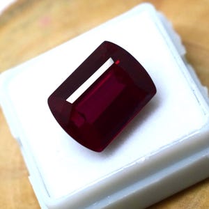 May include: A deep red, rectangular gemstone with faceted edges and a polished surface. The stone features a clear, transparent quality with a distinct light reflection on its top facet, showcasing a rich, saturated crimson hue throughout its body.
