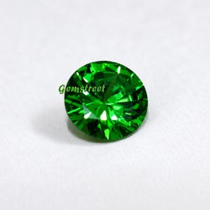 May include: A round, brilliant-cut gemstone with a vibrant, saturated emerald green color. The stone features numerous triangular facets that reflect light, creating a sparkling effect. The text Gemstreet is printed in green on the stone.