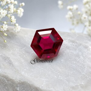 Puede incluir: Una gema de rubí hexagonal y facetada con un tono rojo intenso. La gema está colocada sobre una superficie gris claro, con la palabra "Gemstreet" en escritura cursiva. Flores blancas en el fondo.