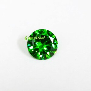 May include: A round, faceted gemstone with a vibrant, saturated green color. The stone features a brilliant cut with numerous triangular facets that reflect light. The word Gemstreet is printed in light green text across the upper left side.