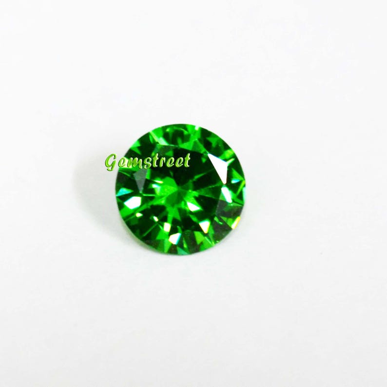 May include: A round, faceted gemstone with a vibrant, saturated green color. The stone features a brilliant cut with numerous triangular facets that reflect light. The word Gemstreet is printed in light green text across the upper left side.