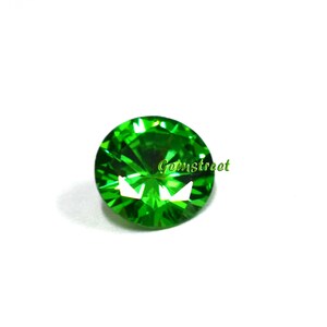 May include: A round, faceted, vibrant green gemstone with a brilliant cut that reflects light in a starburst pattern. The word Gemstreet is printed in a small, light green font across the center of the stone, which has a polished surface.