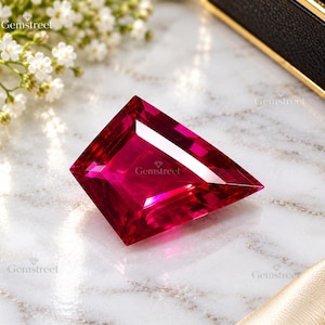 Puede incluir: Una piedra preciosa facetada en forma de escudo de un color rojo magenta intenso y vibrante. La piedra presenta una superficie pulida con bordes geométricos afilados y una claridad transparente brillante que refleja la luz en sus facetas.