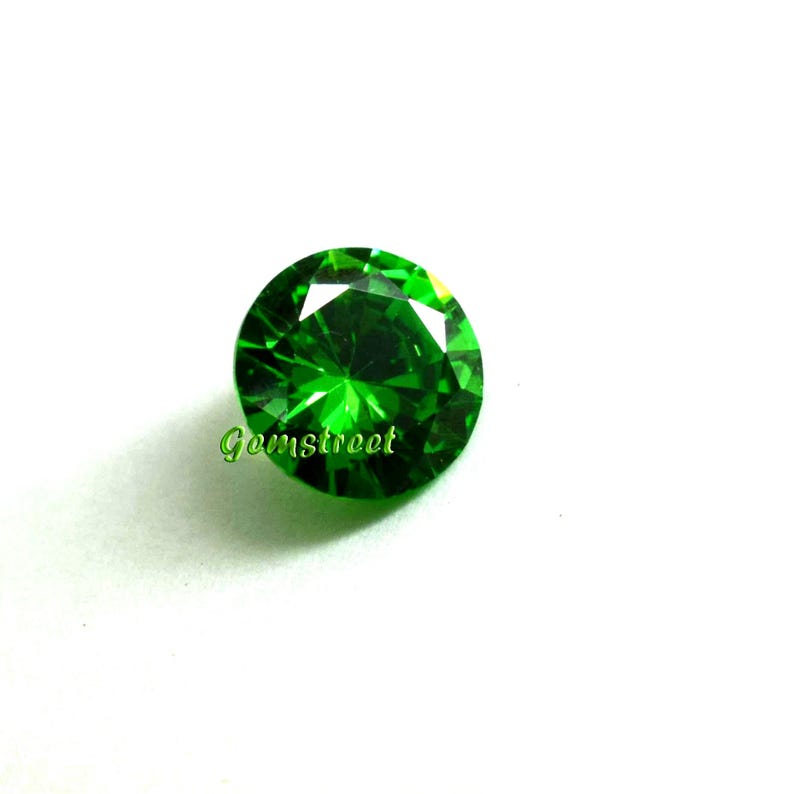 May include: A round, faceted, vibrant green gemstone with a brilliant cut. The stone displays sharp geometric facets that reflect light, creating a sparkling appearance. The word Gemstreet is printed in green text on the lower left side.
