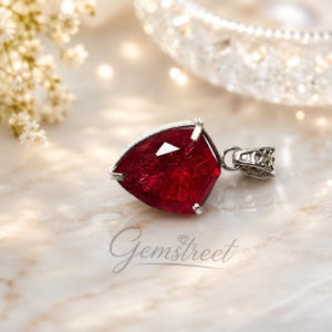 May include: A pendant featuring a faceted, deep red, pear-shaped gemstone set in a silver-toned metal frame with a decorative bail. The word Gemstreet is printed in a cursive font below the pendant, accompanied by a small diamond icon.