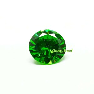 May include: A round, brilliant-cut gemstone with a vibrant, saturated green color and multiple facets that reflect light. The word Gemstreet is printed in a light green, cursive font across the lower right side of the circular stone.