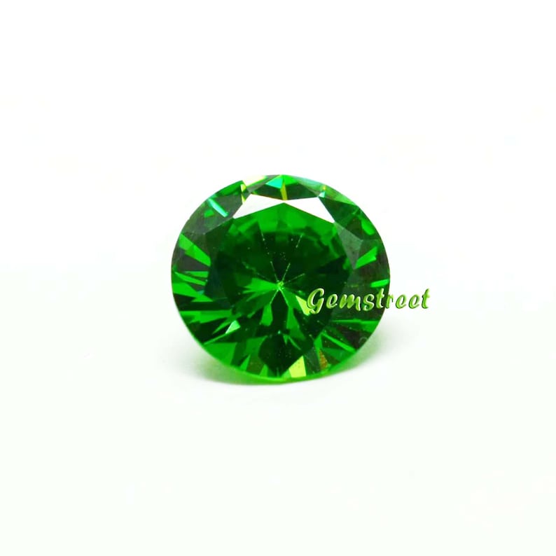 May include: A round, brilliant-cut gemstone with a vibrant, saturated green color and multiple facets that reflect light. The word Gemstreet is printed in a light green, cursive font across the lower right side of the circular stone.