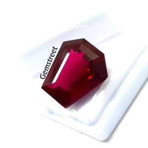May include: A faceted, deep red gemstone with a unique coffin-like shape. The gem is set against a white background, highlighting its rich color and multiple facets. The text "Gemstreet" is printed on the upper left side of the image.