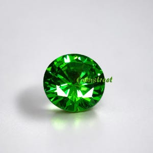 May include: A round, faceted, bright green gemstone. The gem has a brilliant cut, reflecting light. The word "Gemstreet" is written in a cursive font on the gem. The gemstone is set against a white background.