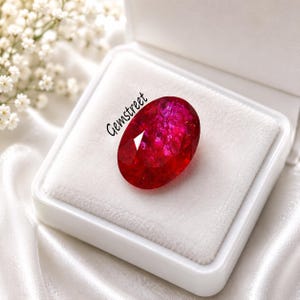 May include: An oval-cut vibrant red gemstone with a faceted surface, featuring the text Gemstreet printed in black. The gemstone is displayed inside a white square jewelry box with a soft white velvet insert, showcasing its deep crimson color.