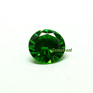 May include: A round, faceted, vibrant green gemstone with a brilliant cut. The stone features a complex geometric pattern of light-reflecting surfaces. The word Gemstreet is printed in green script text on the right side of the gemstone.