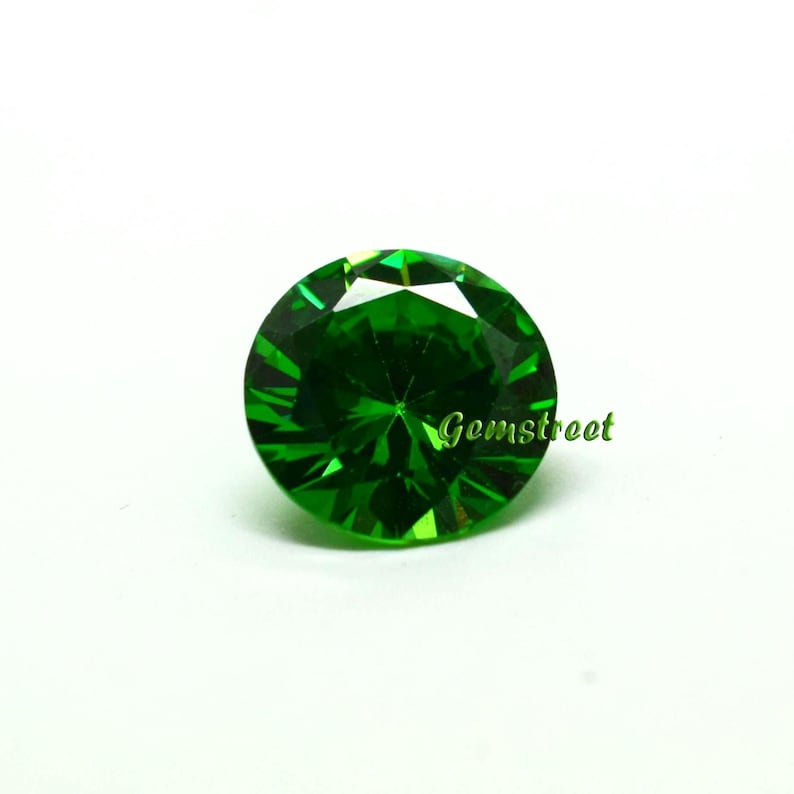 May include: A round, faceted, vibrant green gemstone with a brilliant cut. The stone features a complex geometric pattern of light-reflecting surfaces. The word Gemstreet is printed in green script text on the right side of the gemstone.
