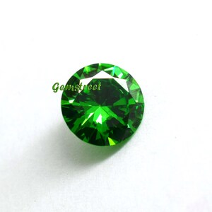 May include: A round, faceted, vibrant green gemstone with a brilliant cut. The stone displays deep emerald hues and sharp geometric light reflections across its surface. The word Gemstreet is printed in green text on the left side of the stone.