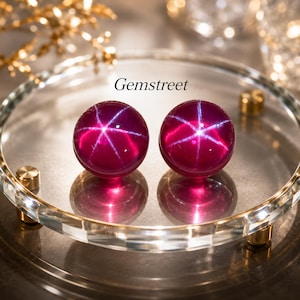 May include: Two round, deep magenta cabochon gemstones featuring a distinct six-rayed star effect on their surfaces. The stones are positioned on a clear glass circular tray. The text Gemstreet is printed in black above the two gemstones.