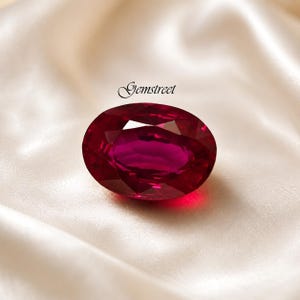 May include: A large, oval-cut ruby gemstone with a deep red hue. The gemstone is set against a white satin background. The word "Gemstreet" is written in cursive above the gemstone.