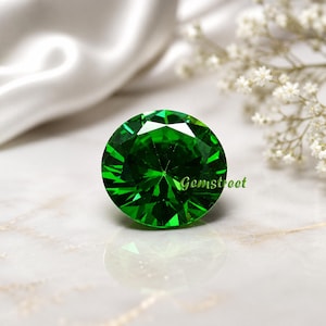 May include: A round, brilliant-cut vibrant green gemstone featuring multiple facets that reflect light. The word Gemstreet is printed in small green letters on the right side of the stone, which displays a clear reflection on the surface below.