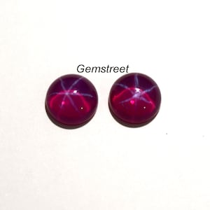 Puede incluir: Dos gemas redondas, pulidas, de color rojo rubí con un efecto de estrella de seis rayos. Las gemas están sobre un fondo blanco. El texto "Gemstreet" es visible.