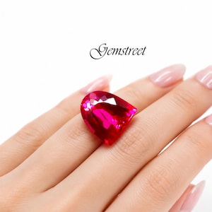 May include: A large, faceted, teardrop-shaped, hot pink gemstone. The gemstone is resting on a hand with long, manicured nails. The word "Gemstreet" is written in cursive above the hand.