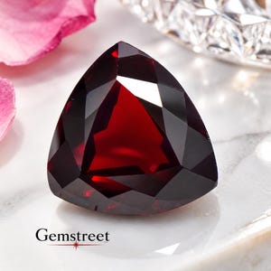 Op de afbeelding: Een dieprode, in trillion-vorm geslepen edelsteen met een gefacetteerd oppervlak dat licht reflecteert. De steen ligt bij roze bloemblaadjes en een helder glazen object. De tekst Gemstreet staat in het zwart linksonder in de afbeelding.