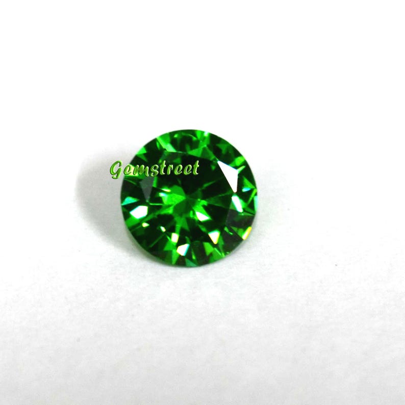 May include: A round, brilliant-cut vibrant green gemstone featuring multiple facets that reflect light. The word Gemstreet is printed in green text on the left side of the stone. The gemstone has a circular shape and a polished surface.