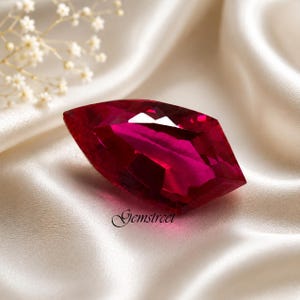Peut inclure: Une pierre précieuse rubis taillée en marquise, d'un rouge profond. La gemme est posée sur un tissu de satin blanc. Le mot "Gemstreet" est écrit en cursive en bas de l'image.