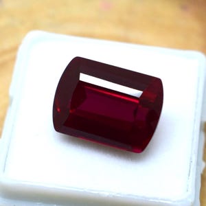 May include: A rectangular, faceted, deep red gemstone. The gem is set against a white background, showcasing its rich color and clarity. The cut of the stone allows light to reflect, creating a brilliant appearance. This gemstone is suitable for jewelry.