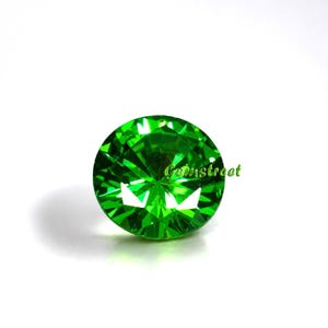 May include: A round, faceted gemstone in a vibrant, brilliant green color. The stone features a precise cut with multiple triangular facets that reflect light, creating a sparkling effect. The text Gemstreet is printed in green on the stone.