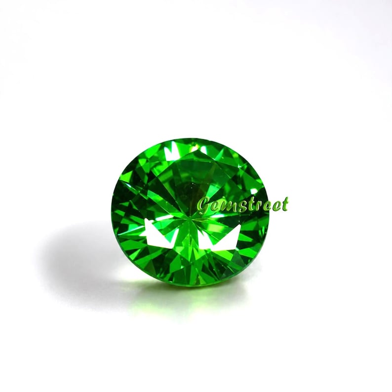 May include: A round, faceted gemstone in a vibrant, brilliant green color. The stone features a precise cut with multiple triangular facets that reflect light, creating a sparkling effect. The text Gemstreet is printed in green on the stone.