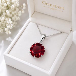 Può includere: Una collana in argento con un ciondolo in pietra preziosa rosso scuro, rotonda e sfaccettata, tenuta da una montatura a griffe. Il gioiello è presentato in una scatola bianca con la scritta Gemstreet stampata in oro sul coperchio interno.