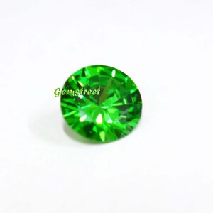 May include: A round, faceted, vibrant green gemstone with a brilliant cut that reflects light. The stone features a clear, intense emerald-like color and is marked with the green text Gemstreet on its surface, showcasing precise geometric facets.