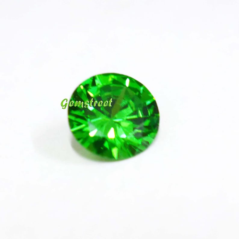 May include: A round, faceted, vibrant green gemstone with a brilliant cut that reflects light. The stone features a clear, intense emerald-like color and is marked with the green text Gemstreet on its surface, showcasing precise geometric facets.