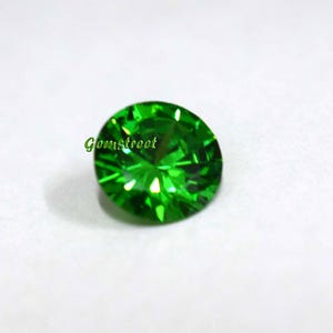May include: A round, faceted, vibrant green gemstone with a brilliant cut. The stone displays sharp geometric light reflections across its surface. The word Gemstreet is printed in green text on the left side of the gemstone's upper surface.