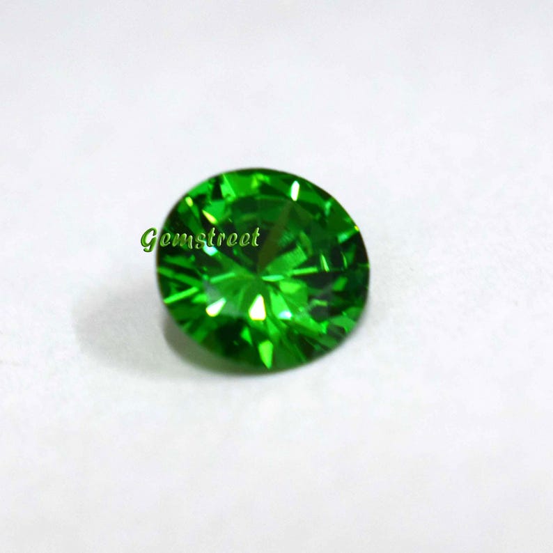 May include: A round, faceted, vibrant green gemstone with a brilliant cut. The stone displays sharp geometric light reflections across its surface. The word Gemstreet is printed in green text on the left side of the gemstone's upper surface.
