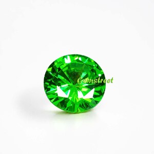 May include: A round, brilliant-cut gemstone with a vibrant, saturated green color and multiple facets that reflect light. The word Gemstreet is printed in a light green font across the center of the stone, which has a clear, polished surface.
