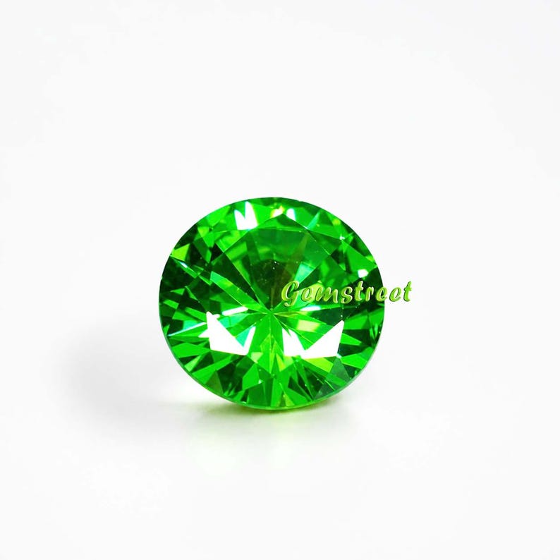 May include: A round, brilliant-cut gemstone with a vibrant, saturated green color and multiple facets that reflect light. The word Gemstreet is printed in a light green font across the center of the stone, which has a clear, polished surface.