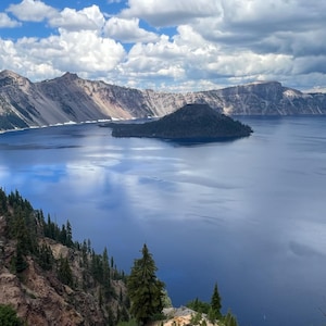 Puede incluir: Un paisaje pintoresco con un gran lago azul profundo rodeado de montañas y acantilados. El cielo está lleno de nubes blancas y esponjosas. Una pequeña isla es visible en el centro del lago, y árboles de hoja perenne salpican el paisaje.