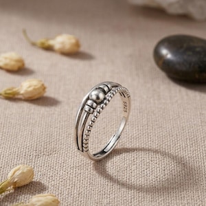 May include: A silver ring featuring a band with a beaded texture on one side and a smooth, polished finish on the other. The ring includes a central spherical bead and two smaller cylindrical accents. The interior of the band is stamped with S925.