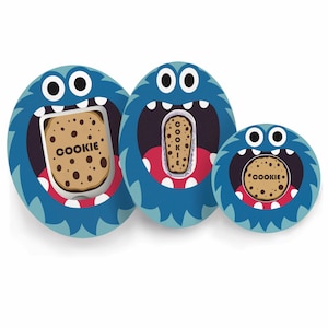 Peut inclure: Trois contenants bleus en forme de monstres avec des yeux blancs et des bouches ouvertes. Chaque monstre tient un biscuit avec le mot "COOKIE" dessus. Les biscuits sont bruns avec des pépites de chocolat. Les contenants sont de tailles différentes.