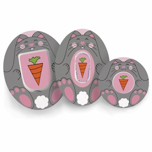 Può includere: Tre ciucci per bambini a forma di coniglietto grigio con dettagli rosa. Ogni ciuccio presenta un centro rosa con un disegno di carota. I coniglietti hanno orecchie, zampe e nasi rosa, con code bianche e soffici.