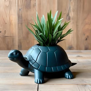 May include: A dark teal turtle-shaped planter with a textured shell design. The planter holds a cluster of green, leafy plants. The turtle has a detailed head, legs, and tail. The planter is on a wooden surface.