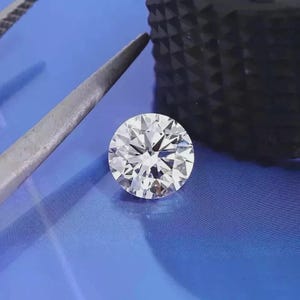 Può includere: Un diamante rotondo con taglio a brillante, con molteplici sfaccettature che riflettono la luce. Il diamante è posto su uno sfondo blu, con uno strumento argentato e un oggetto scuro nel fotogramma. Si sottolinea la purezza e il taglio del diamante.