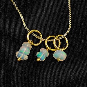 Può includere: Una delicata collana d'oro con tre piccoli pendenti rotondi. Ogni pendente presenta un gruppo di perle opalescenti. La catena della collana e gli anelli dei pendenti sono dorati. Lo sfondo è nero.