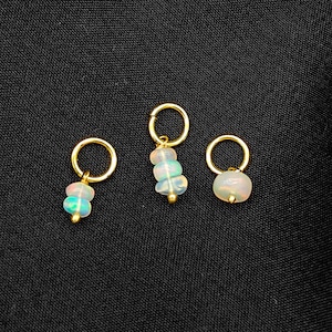 May include: Three gold-colored hoop earrings with opal pendants. The opal pendants have a mix of colors, including white, blue, and pink. The earrings are displayed on a black background.